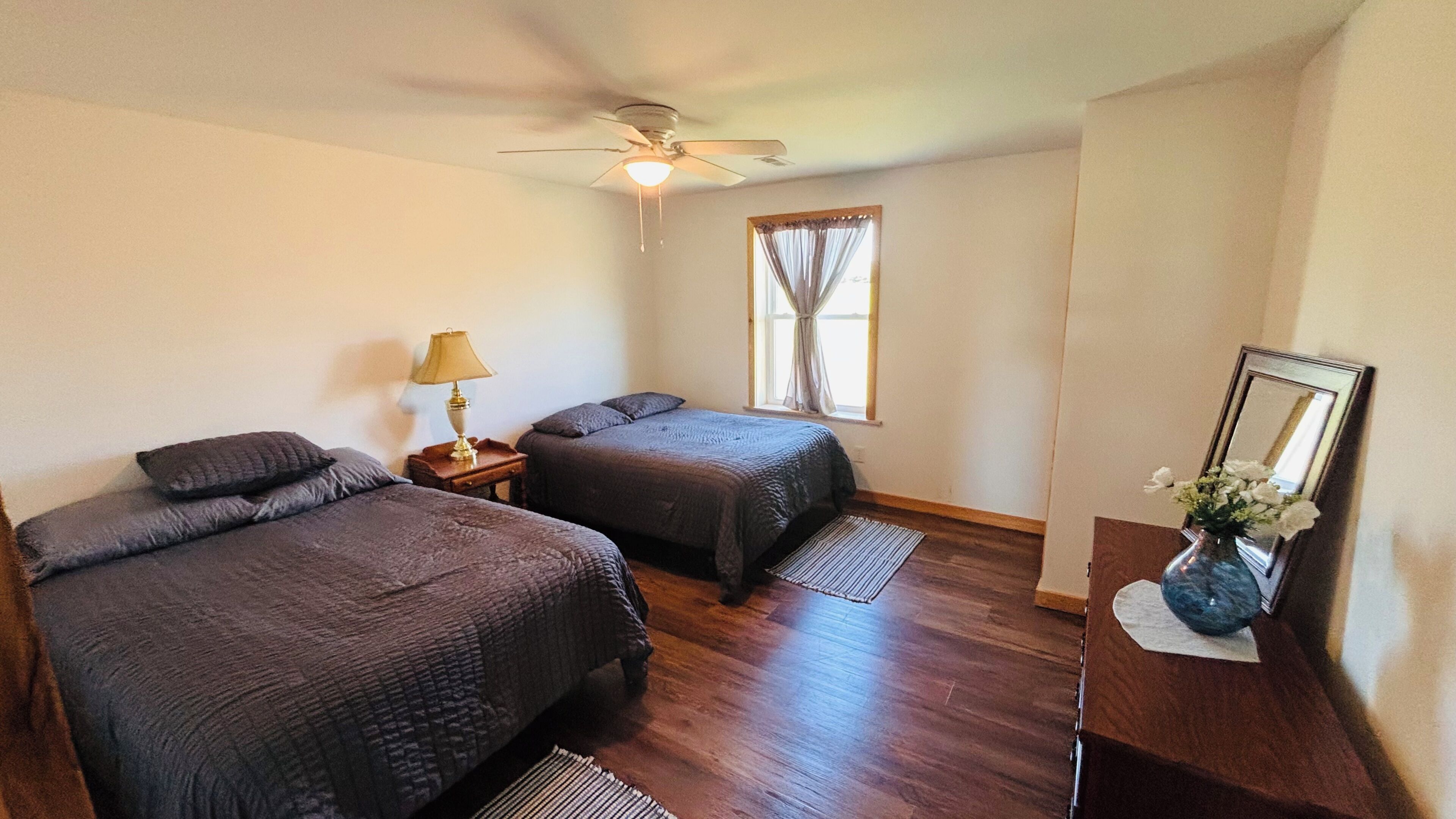A bedroom with two beds, a ceiling fan, a window with curtains, a nightstand with a lamp, and a mirror on a dresser.