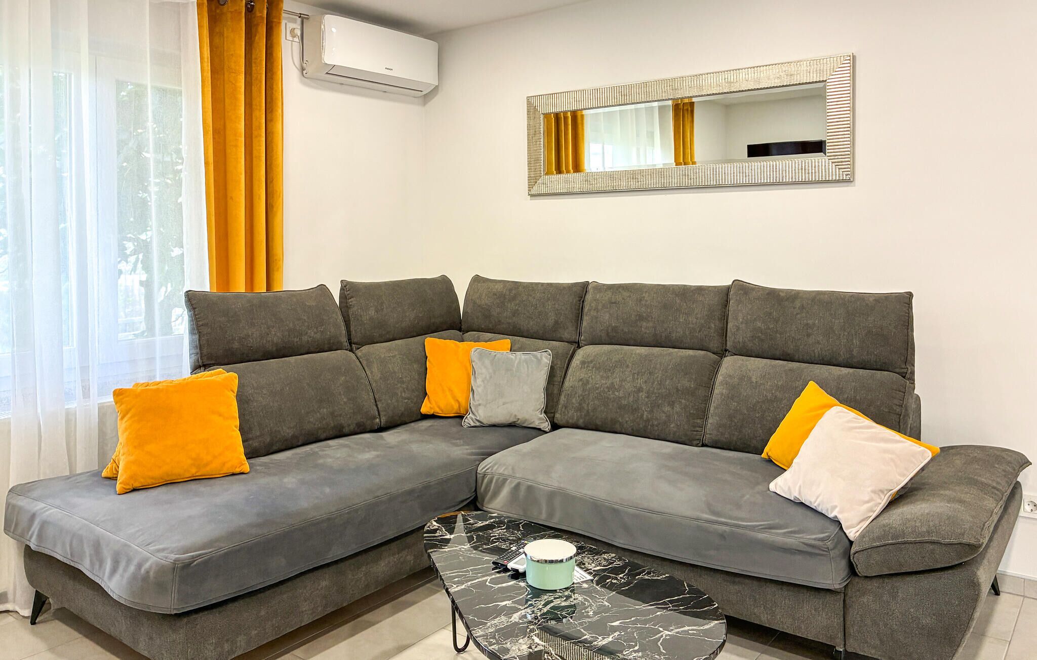 Ein graues Ecksofa mit orangefarbenen und weißen Kissen, ein kleiner schwarzer Marmortisch mit einem weißen Gegenstand sowie eine wandmontierte Klimaanlage.