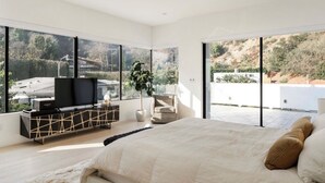 Quarto com uma cama grande, uma TV sobre um console preto e vista para uma encosta.