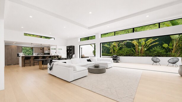 Uma sala de estar moderna com um sofá branco, uma mesa de centro e vista para um jardim verdejante através de grandes janelas.