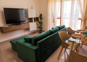 A modern living room with a green sofa, a flat-screen TV, a dining table with chairs, and a potted plant.
