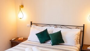 Una cama bien hecha con una colcha a rayas, dos almohadas y toallas enrolladas. Hay una mesita de noche con un libro y un control remoto. Una unidad de aire acondicionado está montada en la pared sobre la cama.