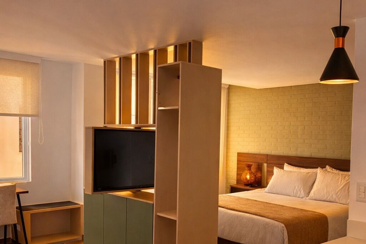 Quarto de hotel compacto com cama, escrivaninha, televisão e cozinha compacta.
