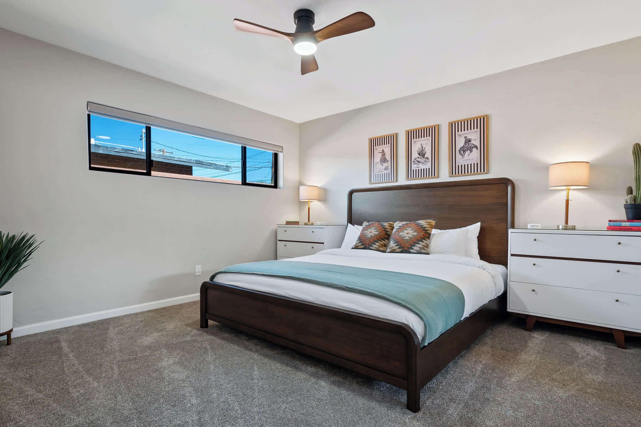 Ein Schlafzimmer mit Bett, Nachttischen, Deckenventilator, einem Fenster mit Blick und Wandbildern.