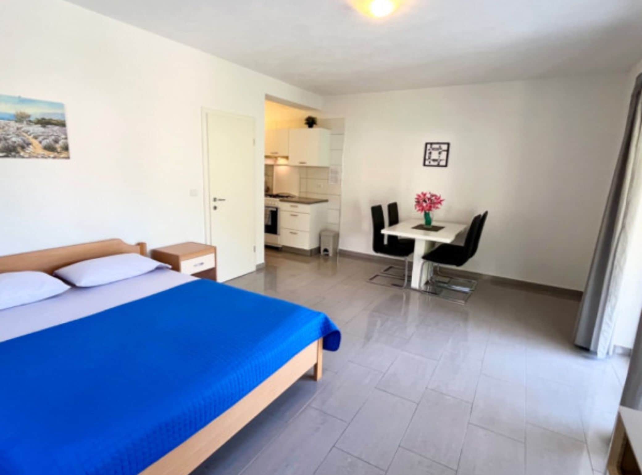Ein Schlafzimmer mit einem Bett, einem kleinen Nachttisch und einem Beistelltisch. Es gibt eine Küchenzeile mit einem Herd, einer Spüle und einer Arbeitsplatte. Ein Essbereich mit Tisch und Stühlen ist sichtbar.