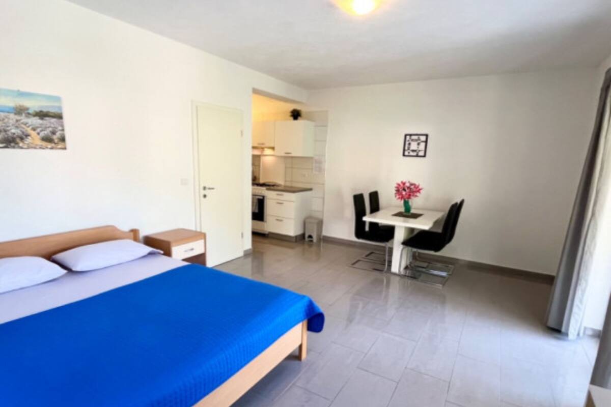 A bedroom with a bed, a small bedside table, and a nightstand. There is a kitchenette with a stove, a sink, and a countertop. A dining area with a table and chairs is visible.