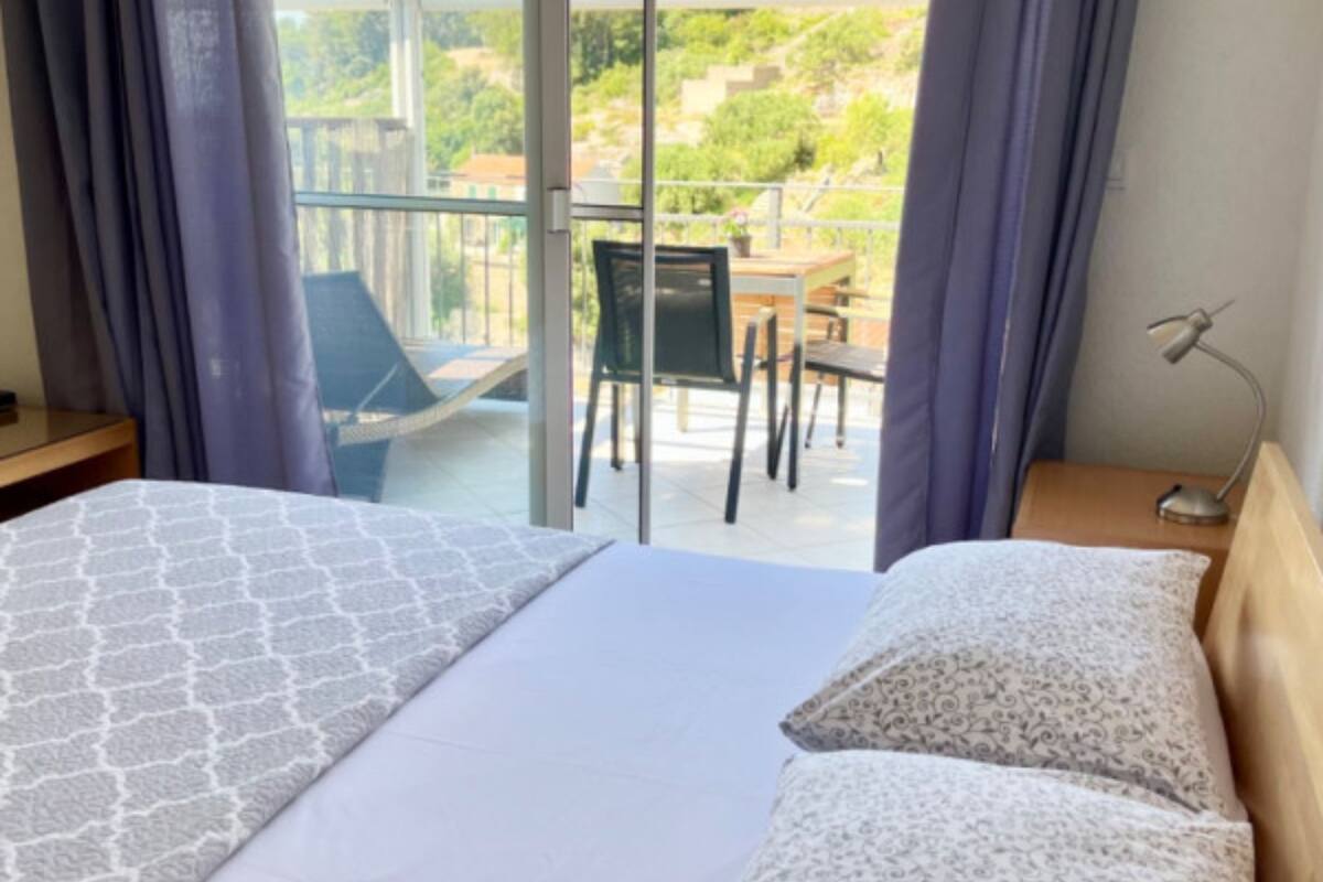 A hotel room with a bed, a chair, a table, and a balcony with a view of greenery.