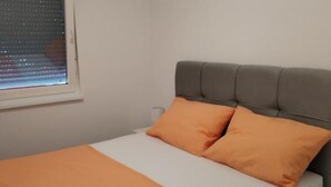 A bed with a grey headboard and two orange pillows, a white mattress, and a window with blinds. - Comfort Apartments Orma (Povljana)