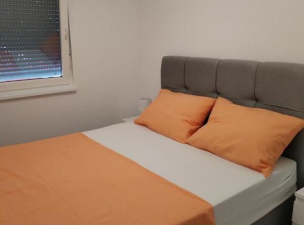Ein Bett mit grauem Kopfteil, zwei orangefarbenen Kissen, einer weißen Matratze und einem Fenster mit Jalousien.