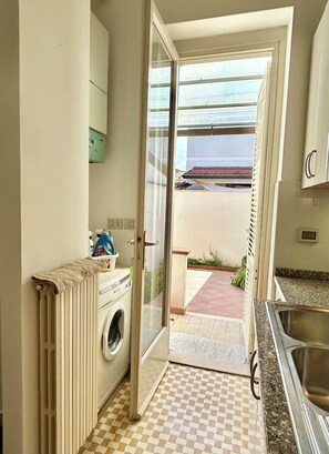 A compact kitchen with a washing machine, a sink, and a view of a beach through an open door. - CasaMilla-Viareggio-villetta-mare-relax-6 places (Viareggio)