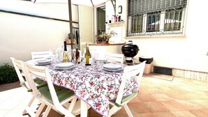 Une table de patio, accompagnée de chaises, d’un barbecue et d’une fenêtre agrémentée d’objets décoratifs, est prête pour un repas en plein air.