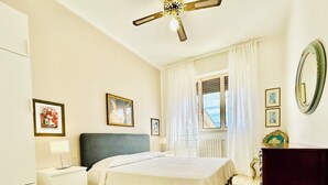 Une chambre à coucher comprenant un lit, une table de chevet, une commode, un ventilateur de plafond, une fenêtre avec des rideaux et un miroir.