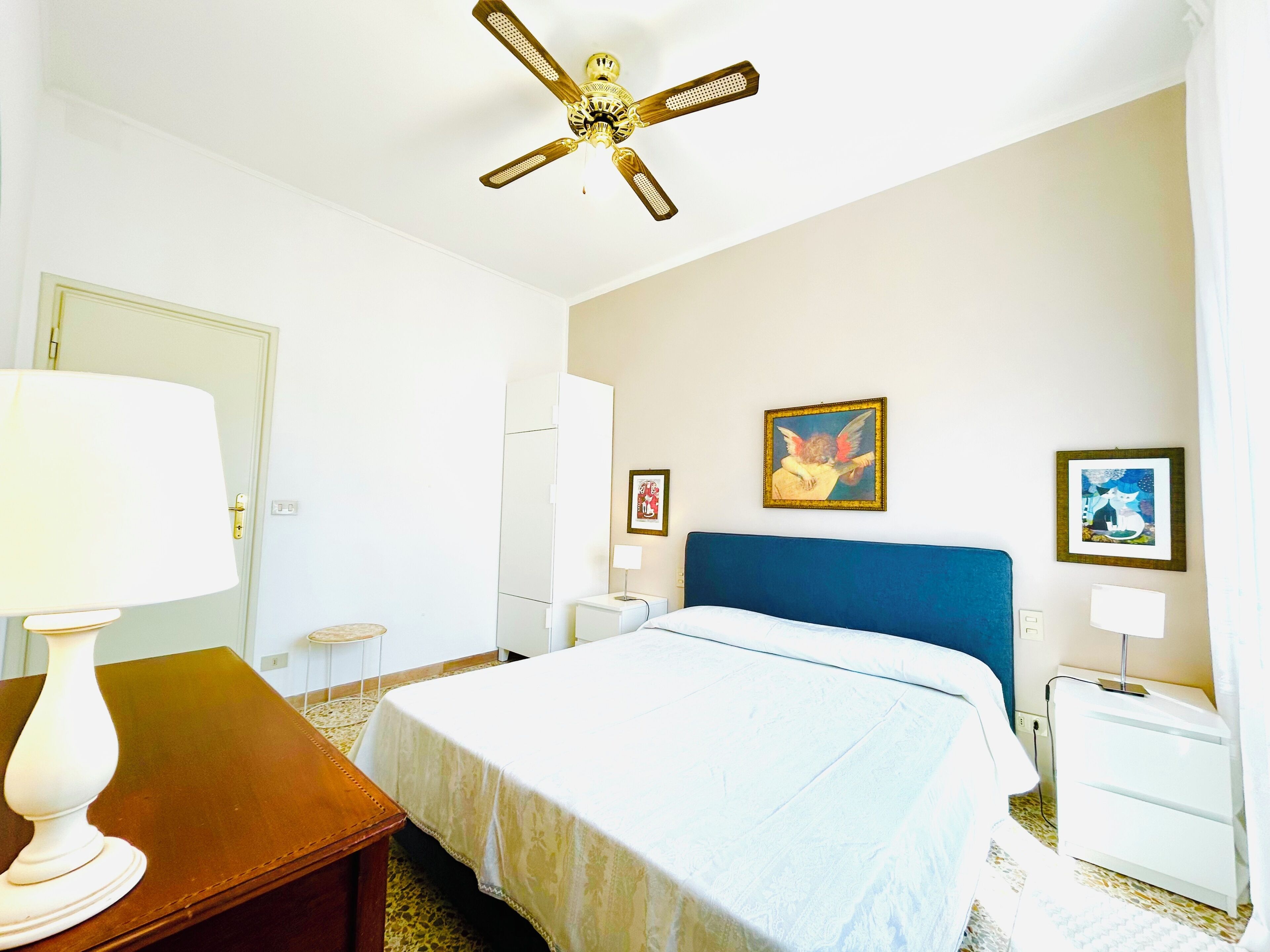 Una habitación de hotel con cama, escritorio, lámpara, ventilador de techo y obras de arte en las paredes.