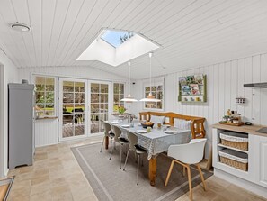 A bright, open-plan kitchen and dining area with a large window, a wooden dining table, chairs, and a built-in shelf with kitchenware.