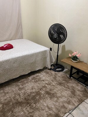 Ein Bett mit weißer Strukturdecke, ein flauschiger beiger Teppich, ein schwarzer Standventilator, ein kleiner Holztisch mit einer Topfpflanze und ein rotes herzförmiges Kissen.