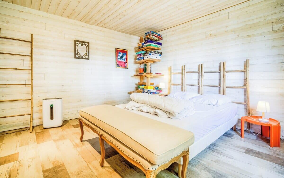 Quarto com cama, um banco, uma escada, uma estante com livros e uma pequena mesa com abajur.