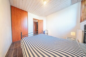 A bedroom with a bed, a chair, a nightstand, a lamp, and a wooden wardrobe. - Pause Vacances : Grand T3 Lumineux (Guidel)