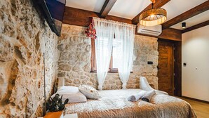 Ein Schlafzimmer mit Steinwänden, einem Bett mit weißen Leinen, einer Holzbalkendecke und einem Fenster mit durchsichtigen Vorhängen.