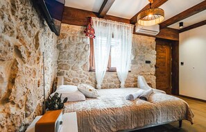 Ein Schlafzimmer mit Steinwänden, einem Bett mit weißen Leinen, einer Holzbalkendecke und einem Fenster mit durchsichtigen Vorhängen.