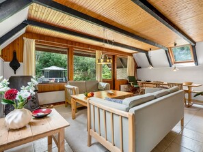 Ein gemütlicher, gut beleuchteter Raum mit Holzbalkendecke, einem Essbereich mit Tisch und Stühlen, einer Couch und einem großen Fenster mit Blick auf die grüne Umgebung.