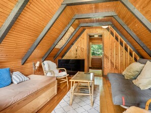 Ein gemütliches Dachzimmer mit Holzbalkendecke, einer Couch, einem Sessel, einem kleinen Tisch und einem Fernseher.