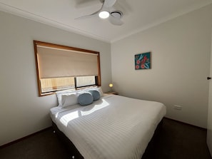 Une chambre à coucher comprenant un lit, un ventilateur de plafond, une fenêtre avec des stores, une lampe de chevet et un tableau accroché au mur.