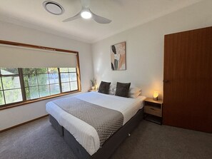 Un dormitorio con cama, mesita de noche, obra de arte colgada en la pared, ventilador de techo y ventana con persianas.