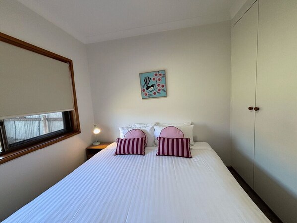 A bedroom with a bed, two pillows, a nightstand, a lamp, a window, a wardrobe, and a wall art piece.