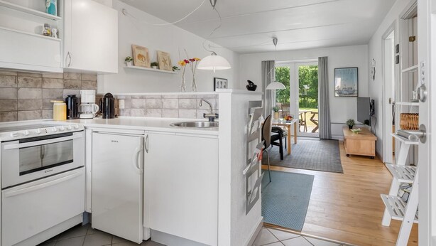 A modern kitchen with white cabinetry, a built-in oven, and a refrigerator. There is a dining area with a table and chairs, and a view of the outdoors through a glass door. - "Enwald" - 100m from the sea by Interhome (Hals)