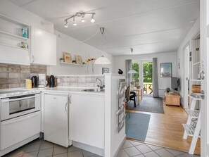 A modern kitchen with white cabinetry, a built-in oven, and a refrigerator. There is a dining area with a table and chairs, and a view of the outdoors through a glass door. - "Enwald" - 100m from the sea by Interhome (Hals)