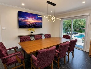 Uma sala de jantar com mesa de madeira e cadeiras vermelhas estofadas, um quadro acima da mesa, e vista para uma piscina através de uma porta de vidro.