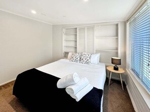 Una habitación de hotel con cama, una mesita de noche con lámpara, una ventana con persianas y estanterías empotradas.