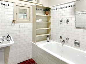 Un baño con paredes de azulejos blancos, un lavamanos blanco, una bañera y un estante con artículos de aseo.