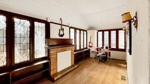 Una habitación con suelos de madera, chimenea de ladrillo, una guitarra, un set de percusión y ventanas amplias con marcos de madera.