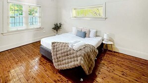 A bedroom with a large bed, two windows with blinds, a bedside table, and a nightstand. - Gurwood Stay - All of the Comforts (Wagga Wagga)