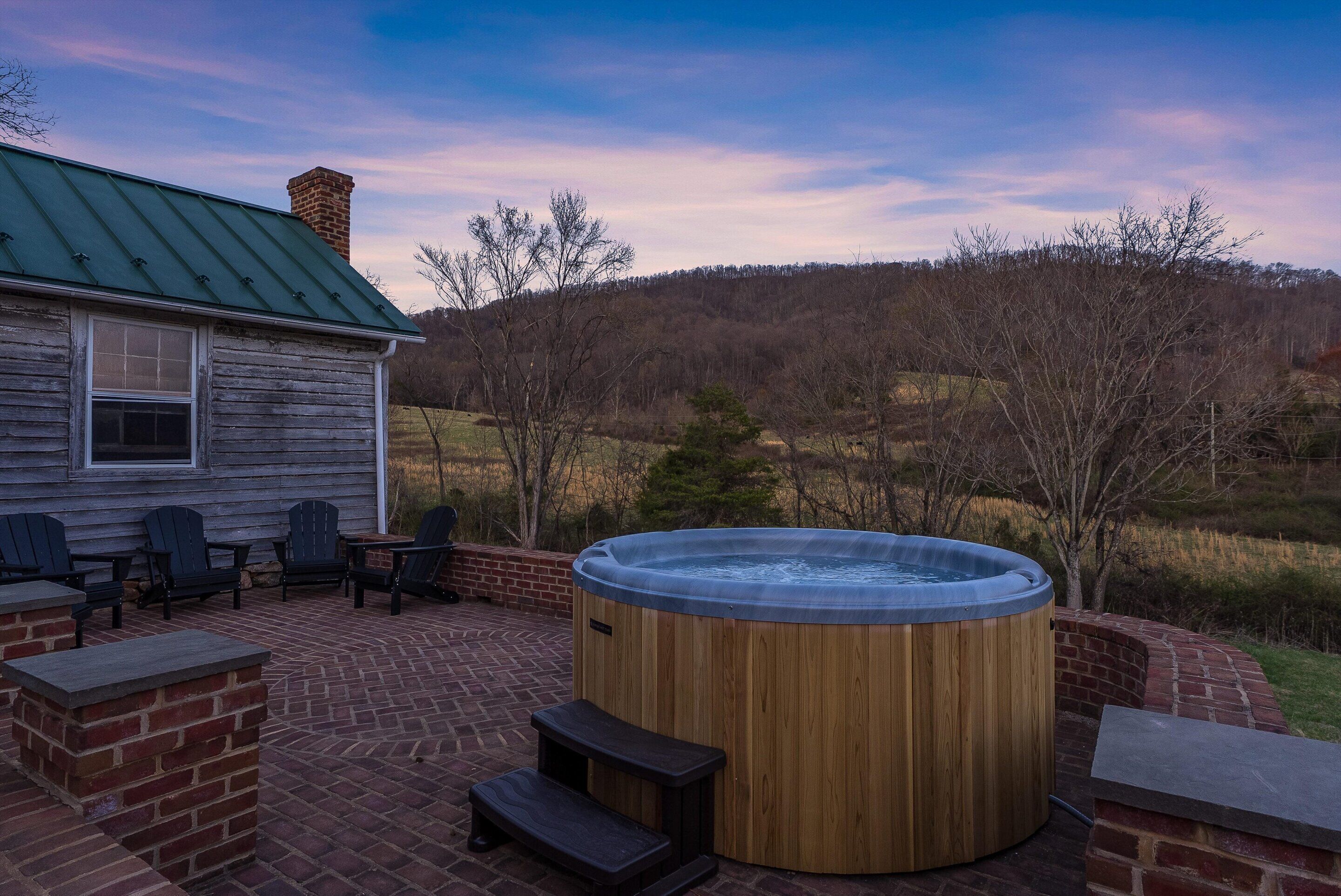 A wooden hot tub with steps, surrounded by brick seating and a patio area with a view of a hilly landscape.