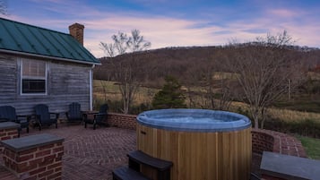 A wooden hot tub with steps, surrounded by brick seating and a patio area with a view of a hilly landscape.