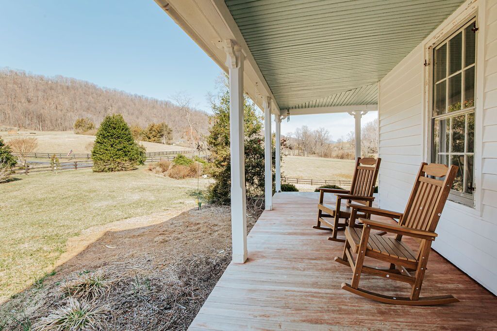 A covered porch with wooden rocking chairs, a white exterior wall, and a view of a grassy field and trees.