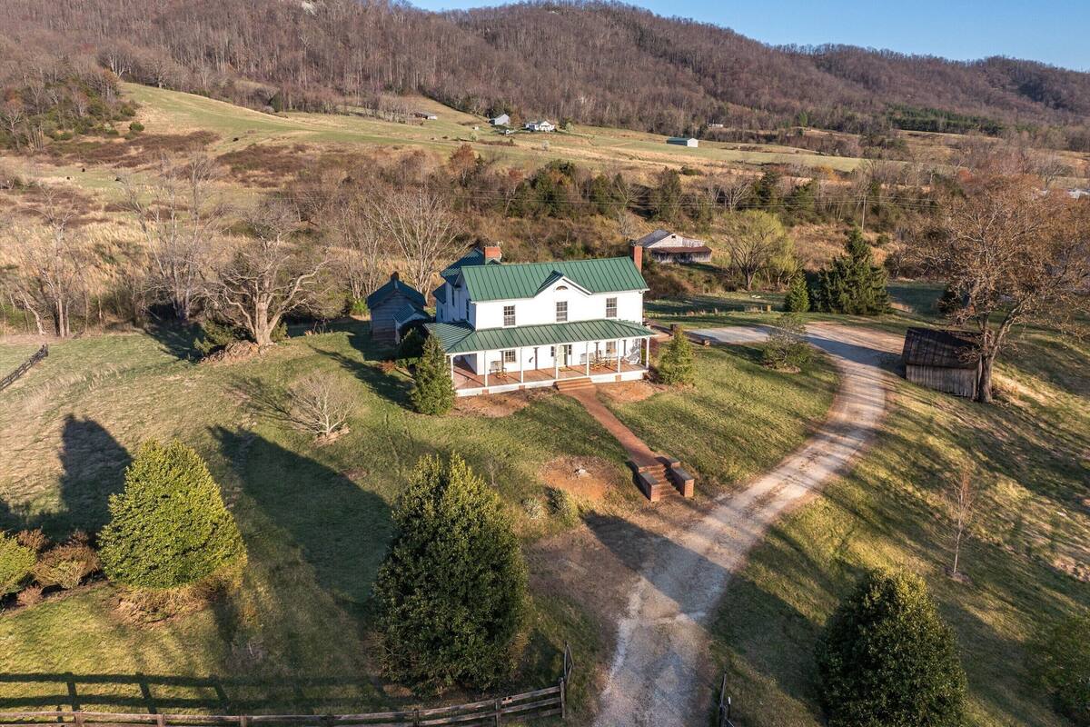 A large house with a green roof, surrounded by a fenced property, set in a hilly rural area.