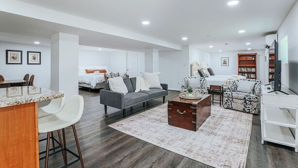 A spacious living area with a sofa, armchairs, a coffee table, and a TV. There is a dining area with chairs and a table. The room has a wooden floor and white walls. - Cavalier Cocoon (Charlottesville)