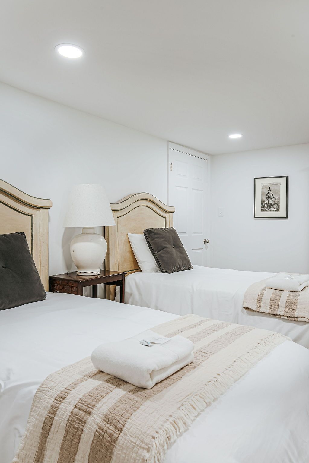 Une chambre d’hôtel avec deux lits, une table de chevet avec une lampe, une porte et un tableau encadré au mur.