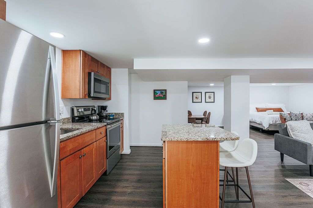 A modern kitchen with stainless steel appliances, wooden cabinets, and a granite countertop. There is a dining area with a table and chairs, and a bedroom with a bed and a gray sofa.