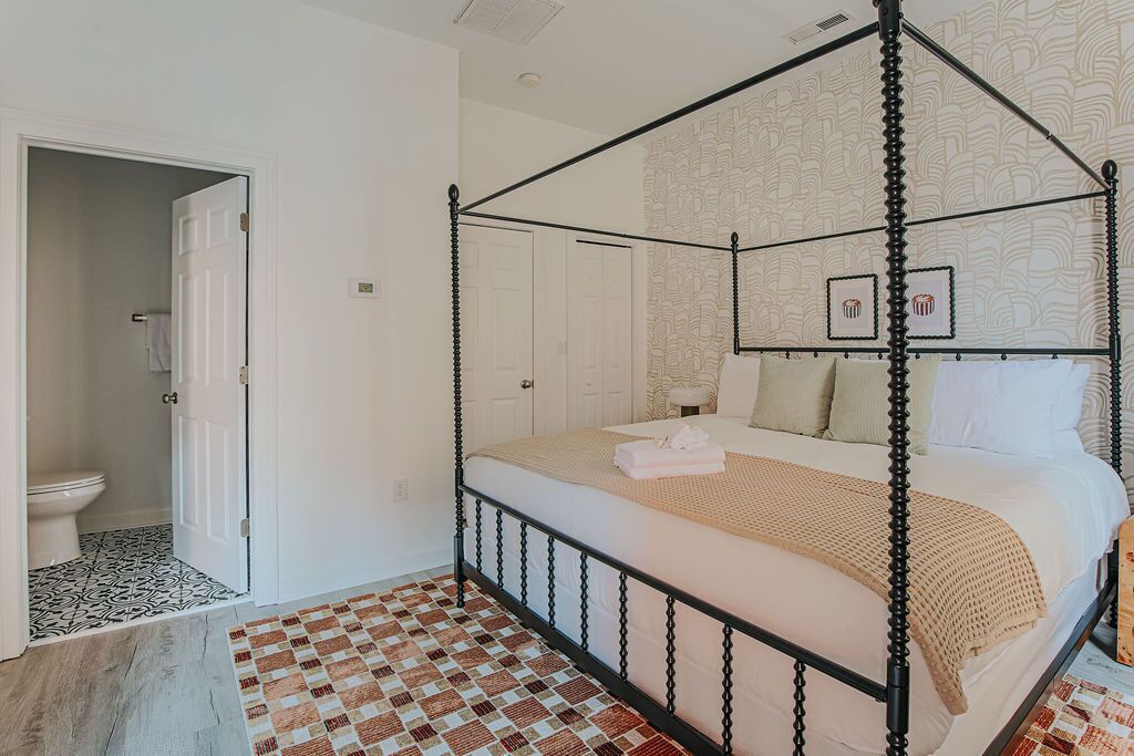 A four-poster bed with a checkered floor, a bathroom with a toilet and patterned tiles, and a door leading to another room.