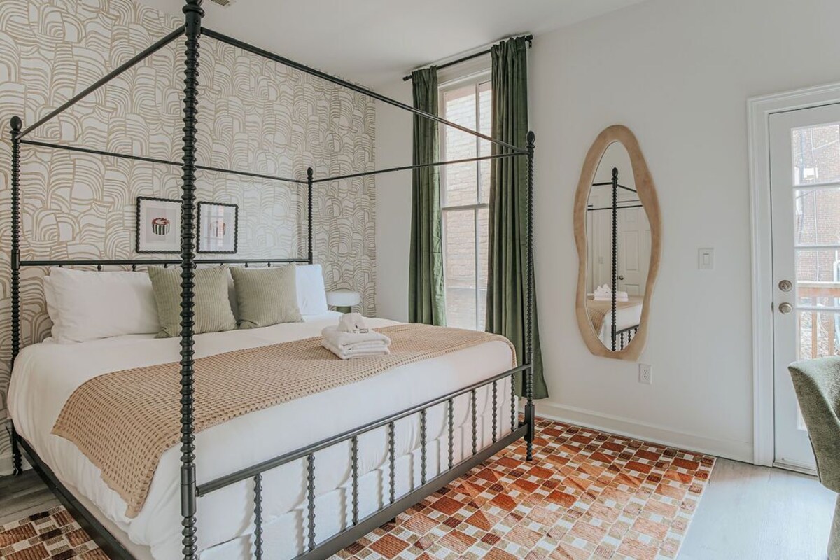 A four-poster bed with a white and beige bedspread, a small bedside table with a lamp, a mirror on the wall, and a checkered floor.