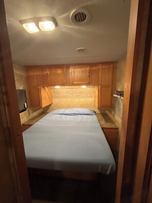 A compact RV interior with a bed, wooden cabinets, and a ceiling vent. -  The SeaNote, Kitty Hawk, NC-- A tranquil stay located just 5 minutes from beach (Kitty Hawk)