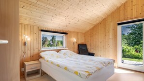 Ein Schlafzimmer mit Holzwandverkleidung, einem Bett, einem Nachttisch und einem Fenster mit Blick auf Grünflächen.