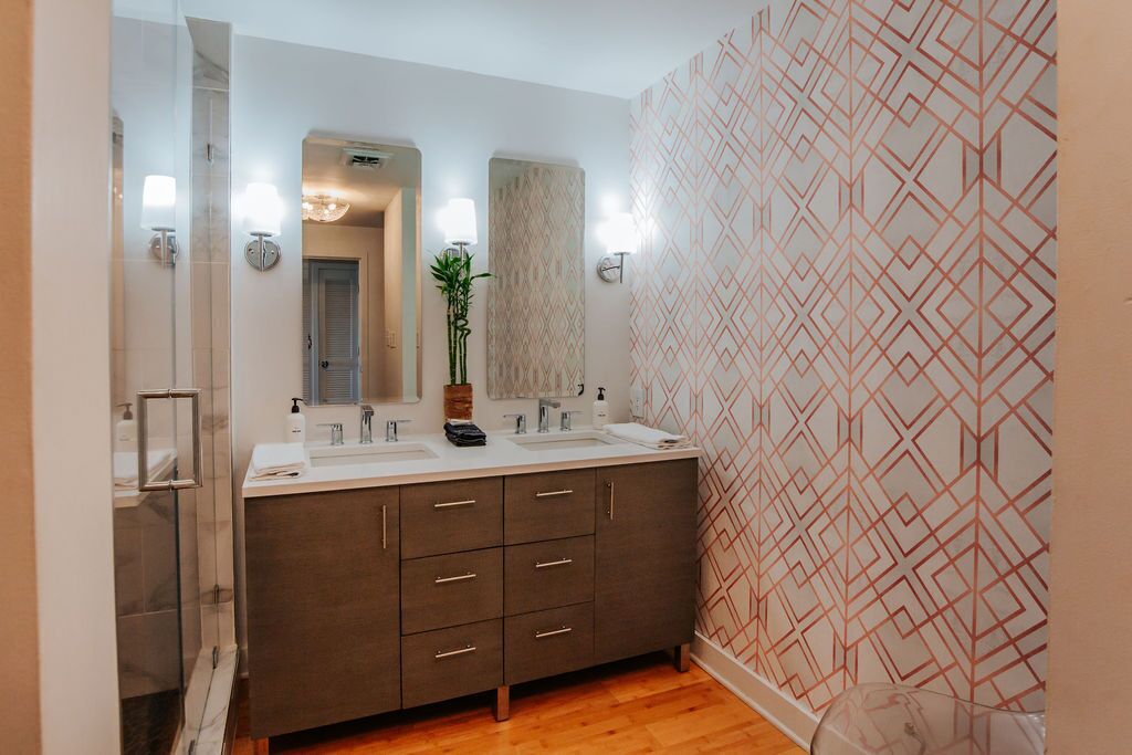 Un baño moderno con tocador doble lavamanos, cabina de ducha de vidrio y pared con patrón geométrico.