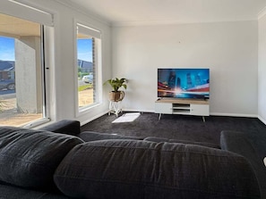 Una sala de estar con un televisor de pantalla plana grande, un sofá gris, una mesita auxiliar con una planta y una ventana que ofrece vistas a un edificio y un estacionamiento.