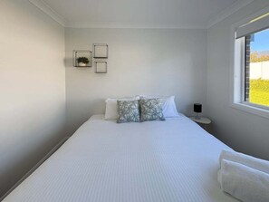 A bedroom with a bed, pillows, a bedside table with a lamp, and a window with a view of greenery.