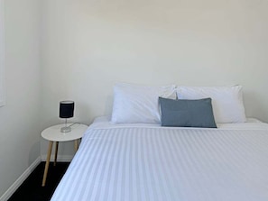 A bed with white bedding and a gray pillow, a small white bedside table with a lamp, and a plain wall.